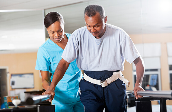 A man is learning to walk using a gait belt and balance beams for assistance with a therapist standing beside